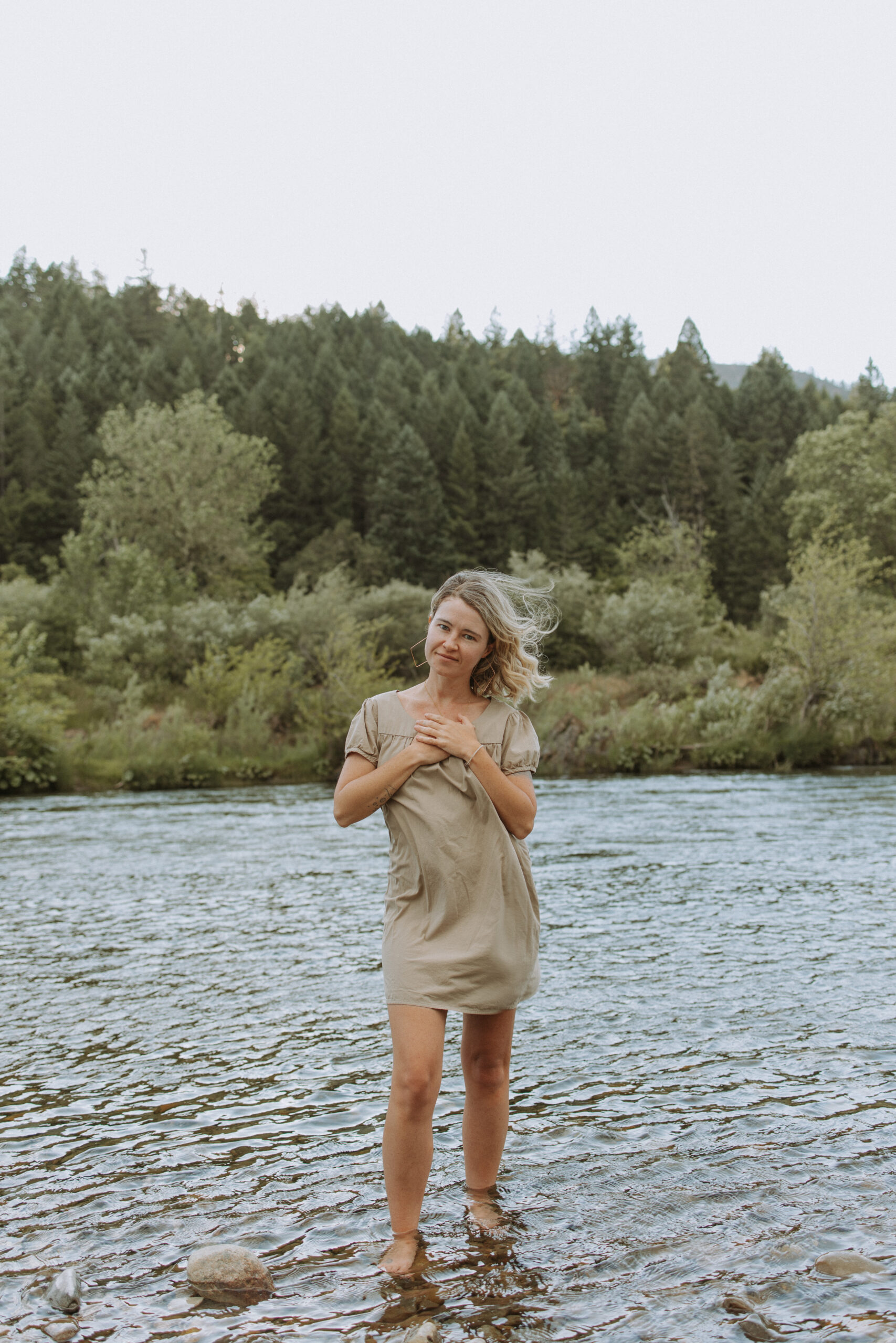 Blonde woman wearing a brown dress holds her hands over her heart smiling. Behind her a river and trees.